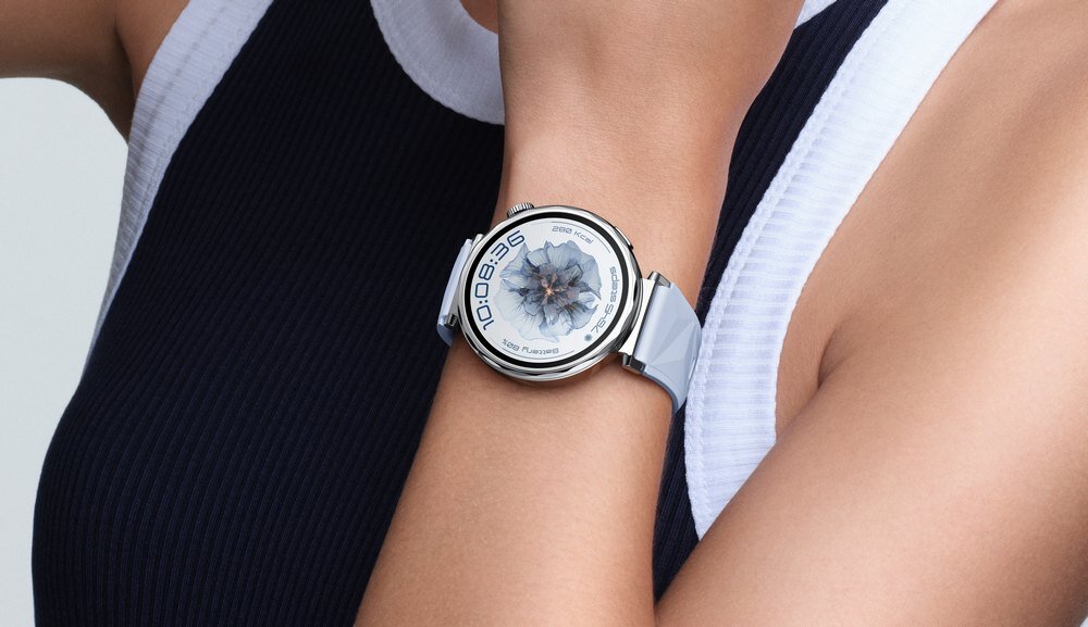Osoba nosi biały smartwatch z okrągłą tarczą, na której widoczny jest cyfrowy wyświetlacz z grafiką przypominającą kwiat. Pasek zegarka jest również biały, a tło stanowi fragment odzieży w ciemnym i jasnym kolorze.