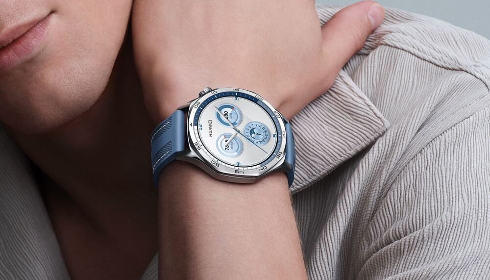 Osoba nosi smartwatch z okrągłą tarczą i niebieskim paskiem. Na tarczy widoczne są trzy mniejsze okręgi oraz logo \'HUAWEI\'.