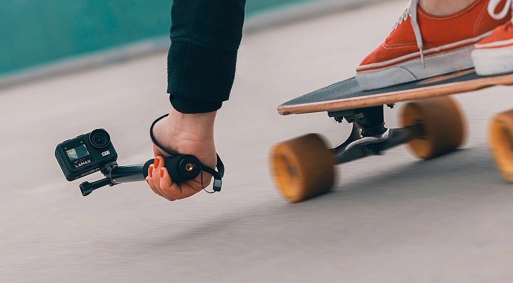 Osoba trzyma kamerę sportową LAMAX W7.1 podczas jazdy na deskorolce. Kamera jest zamocowana na uchwycie, a w tle widać rozmyty obraz podłoża.