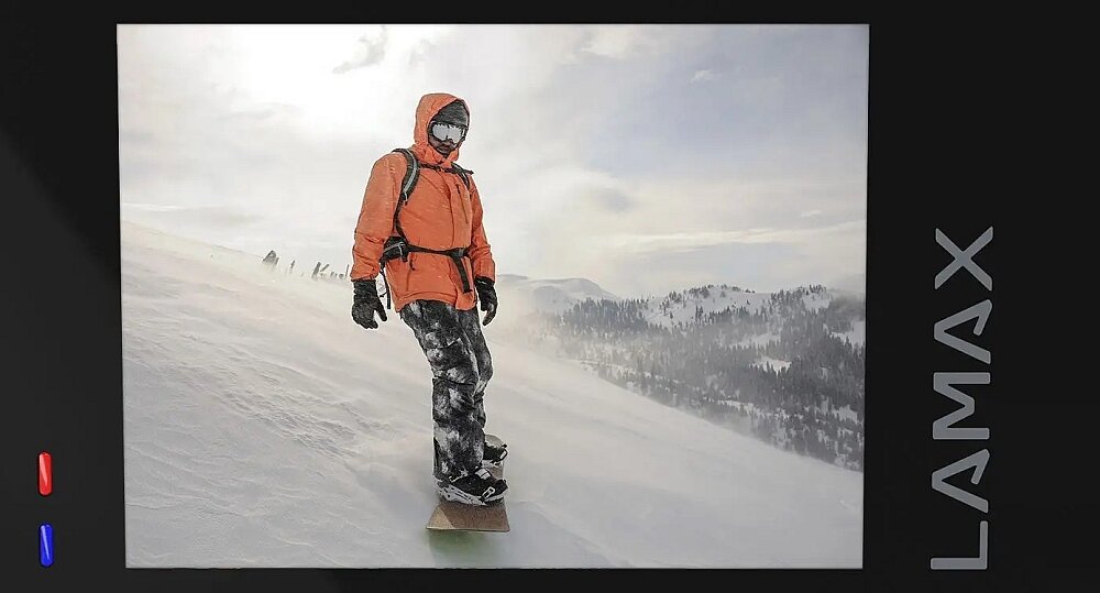 Osoba ubrana w pomarańczową kurtkę i czarne spodnie stoi na desce snowboardowej na zaśnieżonym stoku. Obok zdjęcia widoczne jest logo \'LAMAX\'.