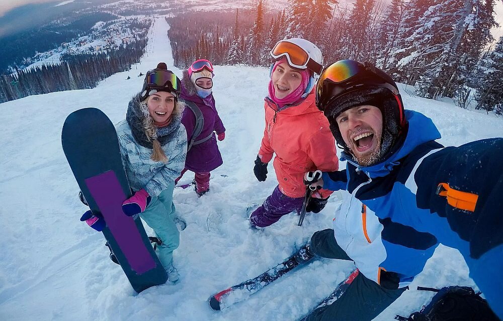Grupa czterech osób w zimowych strojach stoi na zaśnieżonym stoku narciarskim, trzymając sprzęt narciarski i snowboardowy. W tle widać ośnieżone drzewa i rozległy widok na dolinę.