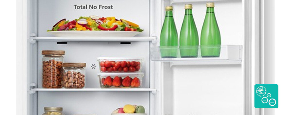 Wnętrze lodówki z napisem \'Total No Frost\', zawierające różnorodne produkty spożywcze, w tym warzywa, owoce, słoiki z orzechami oraz zielone butelki na drzwiach.