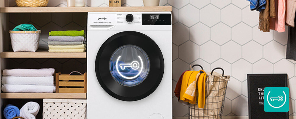 Pralka z logo \'gorenje\' na panelu sterowania, obok kosz z praniem i półki z ręcznikami. Na drzwiach pralki widoczny jest symbol klucza.