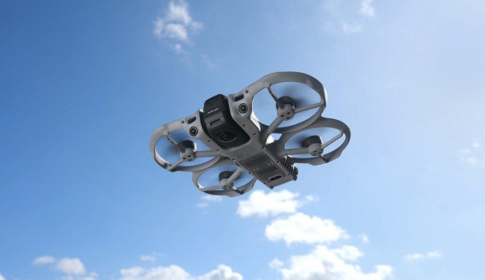 Dron z wbudowaną osłoną śmigieł unosi się na tle nieba z chmurami. Urządzenie ma szary kolor i widoczne są cztery śmigła.