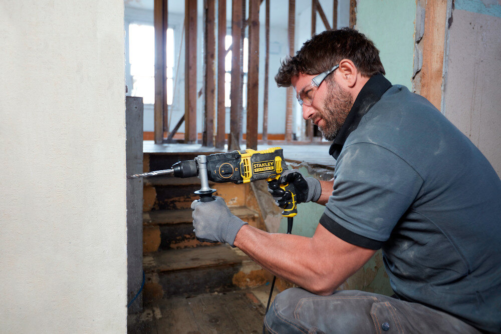 Wiertarka udarowa STANLEY FMEH1100K plac budowy osoba trzyma wiertarkę w dłoniach wierci otwór w ścianie w tle schody drewniane dwubiegowa przekładnia pierwszy bieg z prędkością w zakresie 0-1100 obr/min moment obrotowy do 69 Nm drugi z prędkością 0-3200 obr./ min większa kontrola nad narzędziem precyzyjnie dobrane parametry