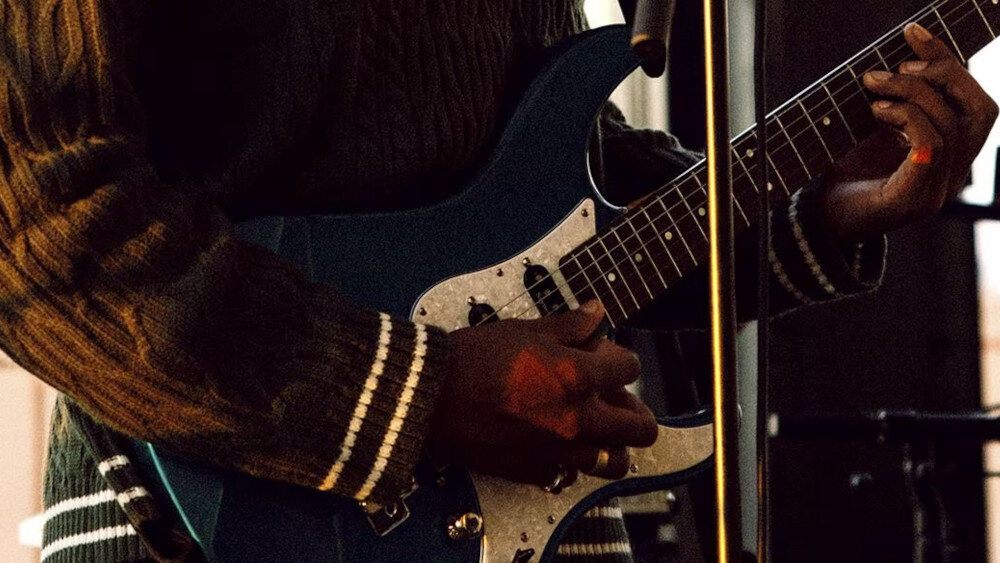 Osoba gra na niebieskiej gitarze elektrycznej, trzymając ją obiema rękami. W tle widoczny jest mikrofon na statywie.