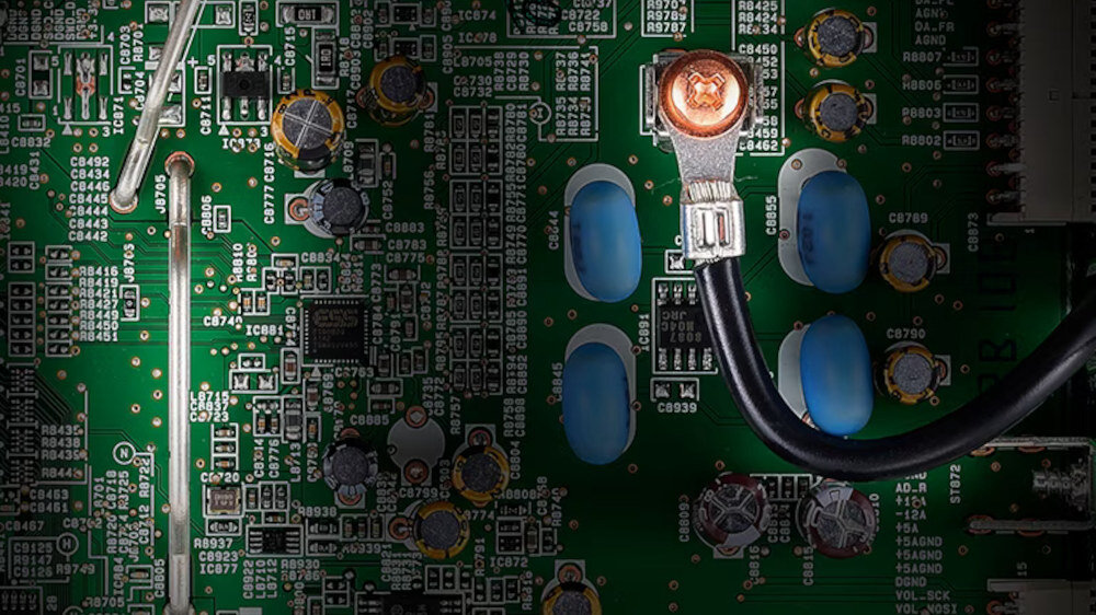 Zdjęcie przedstawia zbliżenie na płytę drukowaną z widocznymi elementami elektronicznymi, takimi jak kondensatory i rezystory. Na płycie znajduje się również czarny przewód przymocowany do metalowego złącza.