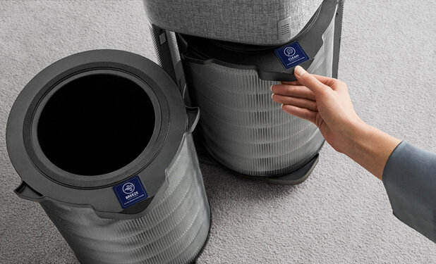 Na obrazie widoczne są dwa cylindryczne filtry do oczyszczacza powietrza z etykietą \'CARE360\'. Ręka dotyka jednego z filtrów, a tło stanowi szara wykładzina.
