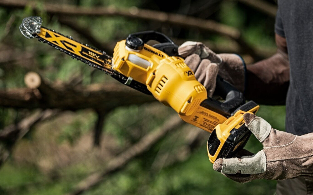 Osoba trzyma żółtą piłę łańcuchową z widocznym logo DEWALT i oznaczeniem 18V. W tle znajdują się rozmyte gałęzie drzew.