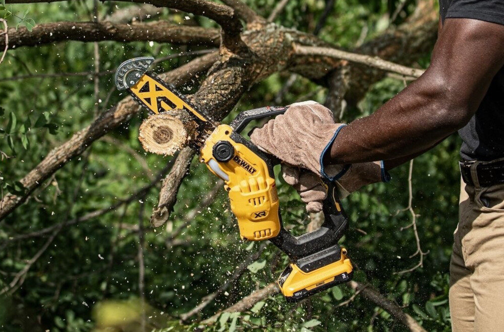 Osoba używa żółtej piły łańcuchowej do cięcia gałęzi drzewa. Na piłę widoczne jest logo \'DEWALT\'.