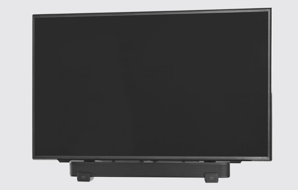 Na obrazie widoczny jest telewizor z zamontowanym poniżej soundbarem w kolorze czarnym. Soundbar jest przymocowany za pomocą uchwytu, a na jego końcach znajdują się dwa prostokątne elementy.