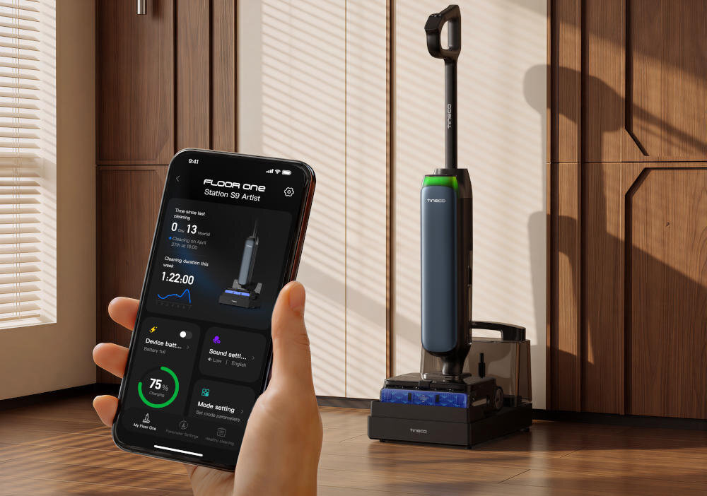 Na obrazie widoczny jest odkurzacz stojący na stacji dokującej oraz ręka trzymająca smartfon z aplikacją pokazującą dane dotyczące urządzenia. Na ekranie telefonu widoczny jest tekst: \'FLOOR One Station S9 Artist\'.