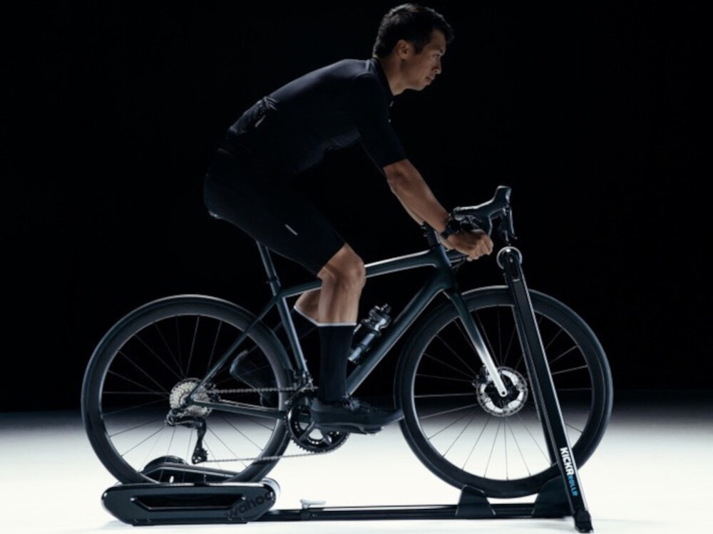 Osoba na rowerze korzysta z trenażera rowerowego w ciemnym otoczeniu. Na trenażerze widoczne jest logo \'KICKR\'.