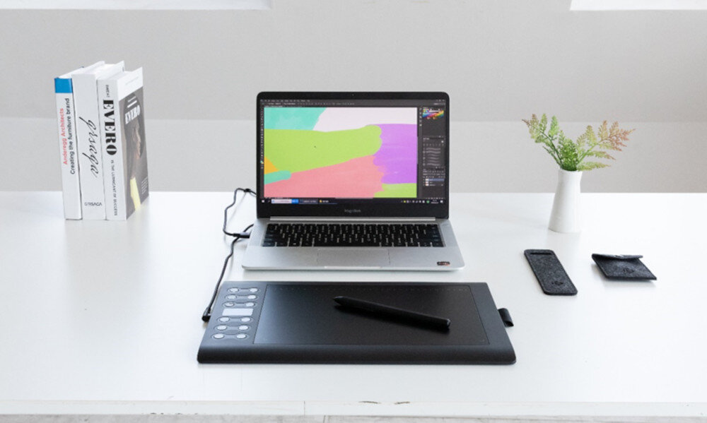 Na białym biurku znajduje się laptop z kolorowym projektem na ekranie, obok leży czarny tablet graficzny z piórem. W tle widać kilka książek i małą roślinę w białej doniczce.