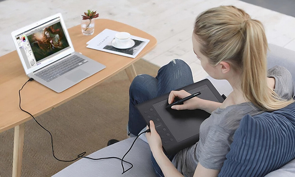 Osoba siedzi na kanapie, używając tabletu graficznego podłączonego do laptopa, na którym wyświetlany jest program graficzny. Na stoliku obok znajduje się filiżanka z napojem i mała roślina w doniczce.