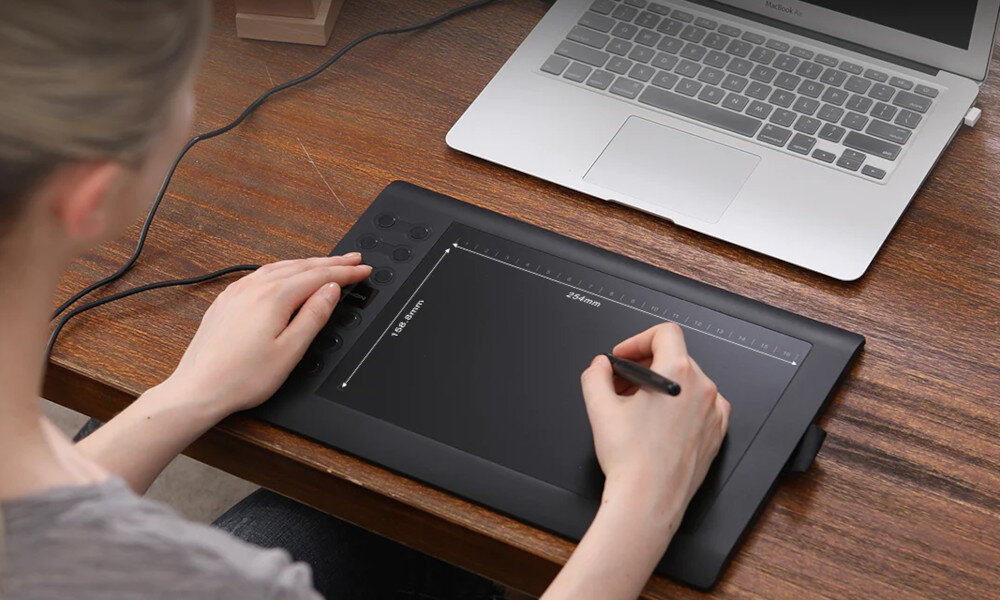 Osoba używa czarnego tabletu graficznego z piórem, obok znajduje się otwarty laptop na drewnianym biurku.