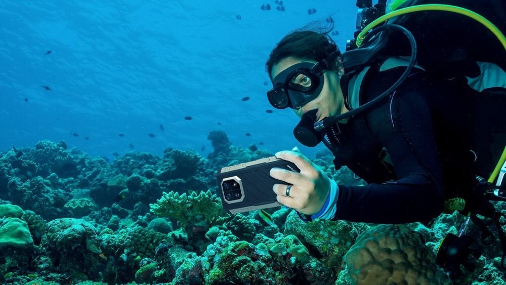 Osoba w stroju do nurkowania trzyma smartfon pod wodą, otoczona rafą koralową i rybami. Smartfon ma solidną obudowę, a w tle widać błękitną wodę.