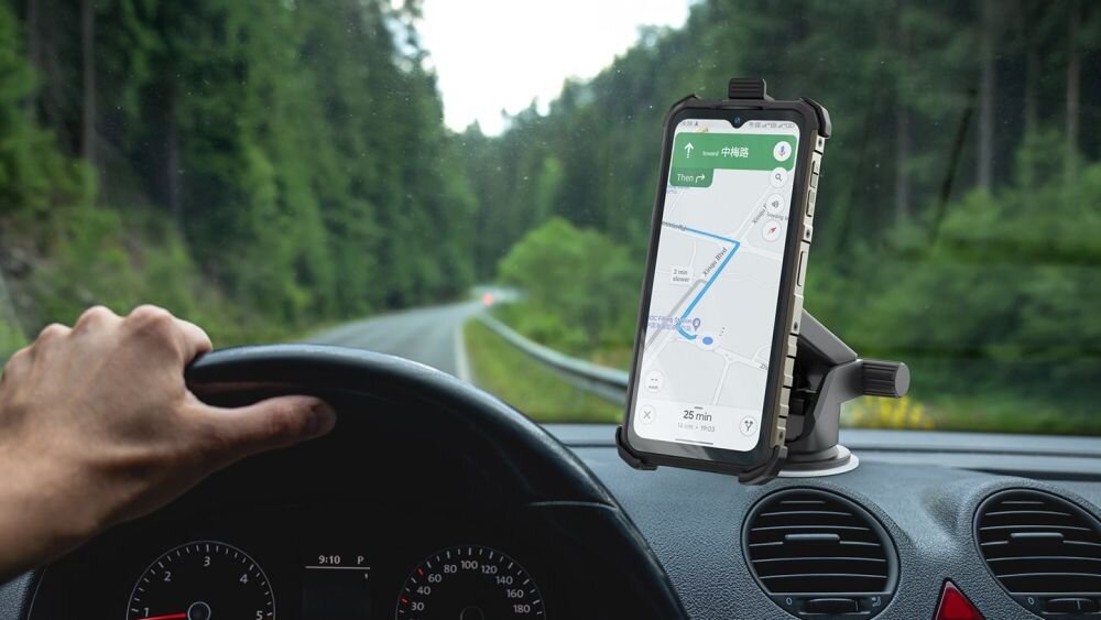 Smartfon zamocowany na desce rozdzielczej samochodu wyświetla mapę z trasą. W tle widoczna jest droga otoczona drzewami.