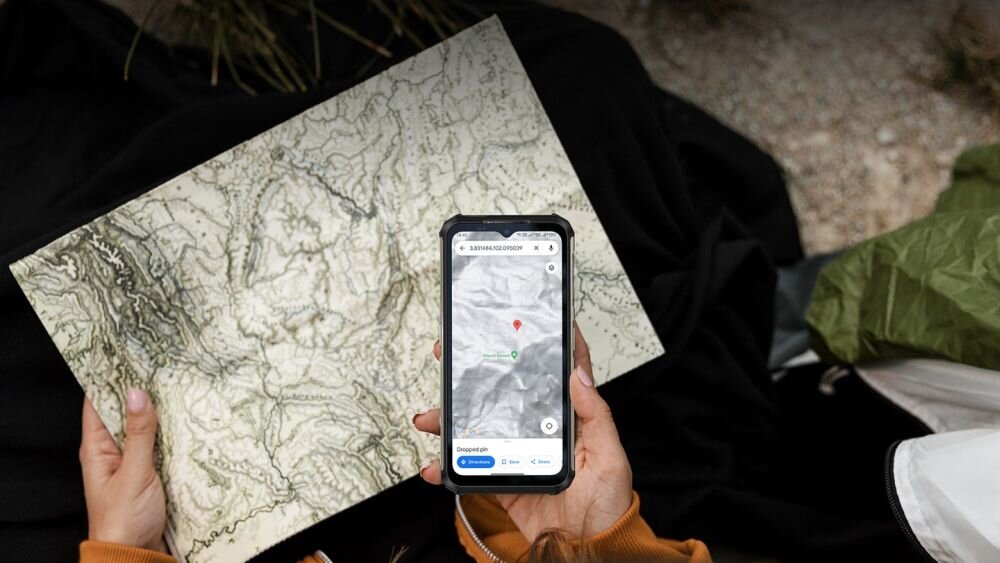 Osoba trzyma smartfon z wyświetloną mapą oraz papierową mapę topograficzną. Na ekranie telefonu widoczny jest punkt oznaczony pinezką na mapie.