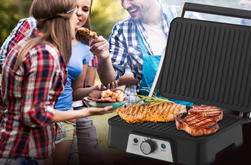 Grill elektryczny z otwartą pokrywą, na którym znajdują się grillowane kawałki mięsa i warzywa. W tle widoczni są ludzie trzymający talerze z jedzeniem.