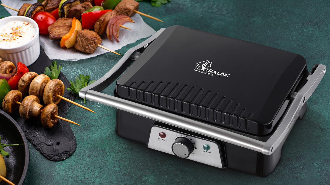 Na obrazie znajduje się czarny grill elektryczny z logo \'EXTRALINK\' na pokrywie. Obok grilla leżą szaszłyki z warzywami i mięsem oraz miseczka z sosem.