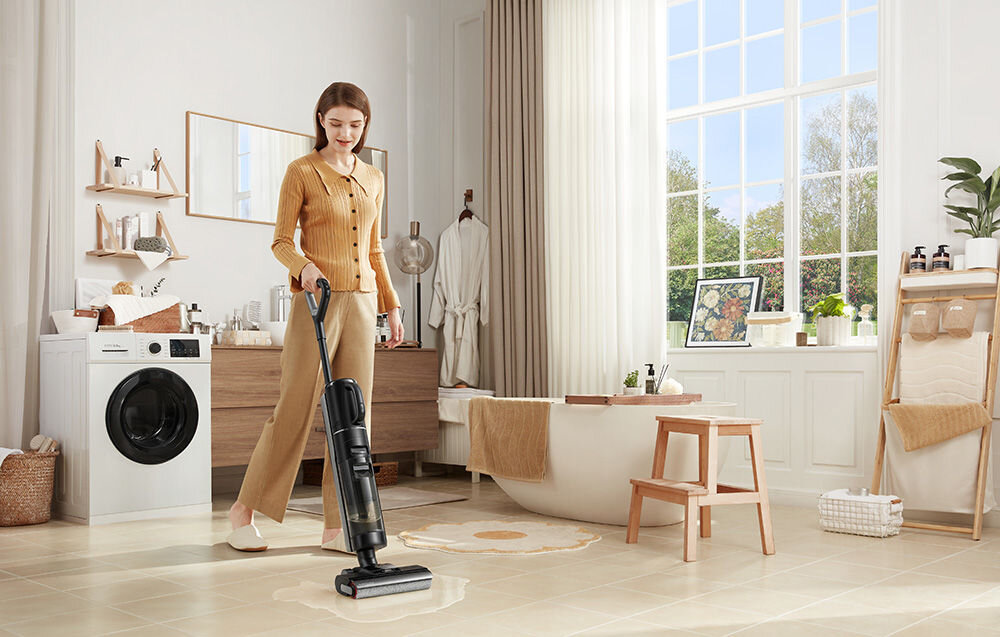 Kobieta używa odkurzacza mopującego na jasnej podłodze w eleganckiej łazience. W tle widoczna jest pralka, wanna oraz duże okno z widokiem na zieleń.