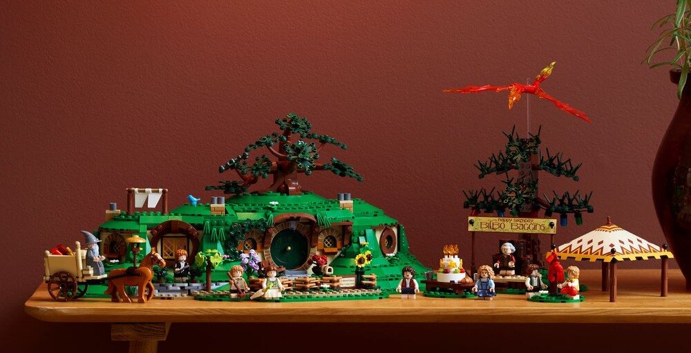 Na drewnianym stole znajduje się zestaw LEGO przedstawiający scenę z zielonym wzgórzem, domkami wbudowanymi w ziemię, drzewami i postaciami. W tle widoczny jest czerwony smok, a na jednym z elementów napis \'Frodo Baggins\'.