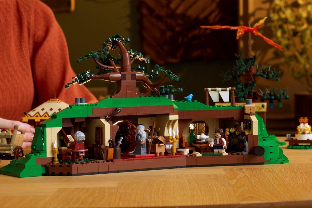 Zestaw LEGO przedstawiający wnętrze domu z postaciami i szczegółami, takimi jak meble i dekoracje. W tle widoczna jest osoba oraz elementy krajobrazu z drzewem i latającym stworzeniem.