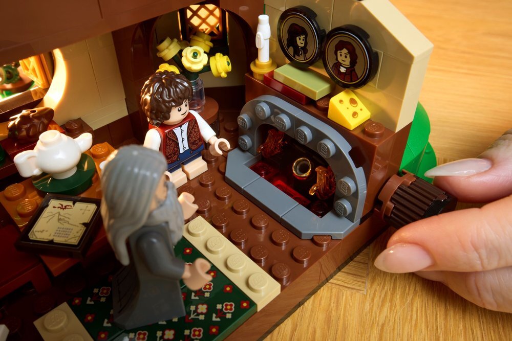 Zestaw LEGO przedstawiający wnętrze domu z kominkiem, przy którym znajdują się dwie figurki. W tle widoczne są kwiaty, obrazki na ścianie oraz ręka obracająca element konstrukcji.