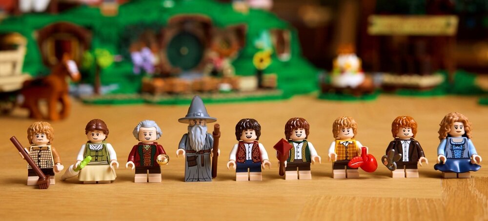Na drewnianym stole ustawiono osiem figurek LEGO przedstawiających postacie z filmu. W tle widoczna jest makieta zielonego wzgórza z okrągłymi drzwiami i kwiatami.