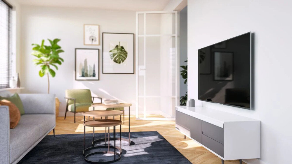 Nowoczesny salon z telewizorem zamontowanym na ścianie, obok białej szafki. W pomieszczeniu znajdują się również sofa, stoliki kawowe i rośliny doniczkowe.