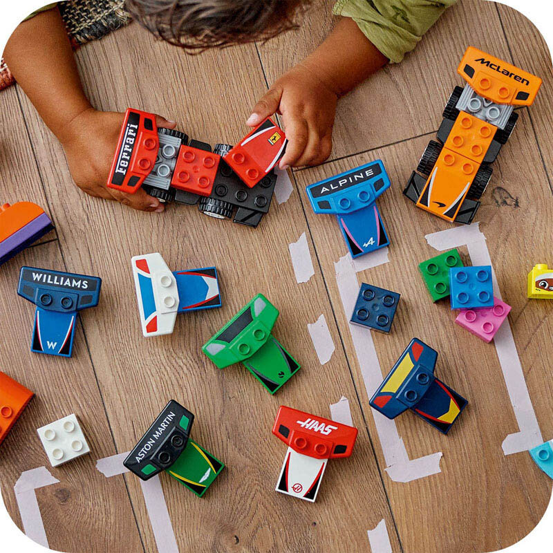 Na drewnianej podłodze znajdują się kolorowe klocki LEGO w kształcie bolidów z widocznymi nazwami: Ferrari, McLaren, Williams, Alpine, Aston Martin, Haas. Dziecko bawi się klockami, a obok leżą dodatkowe elementy w różnych kolorach.
