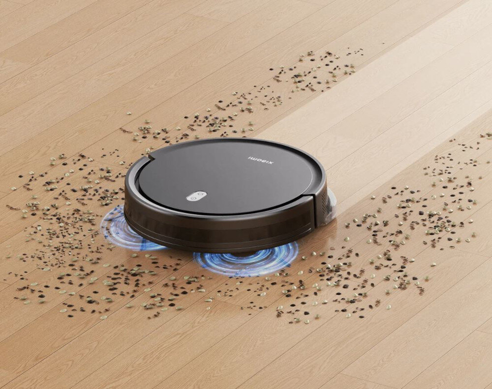 Robot sprzątający w kolorze czarnym porusza się po drewnianej podłodze, zbierając rozsypane drobinki. Na urządzeniu widoczne jest logo \'Xiaomi\'.