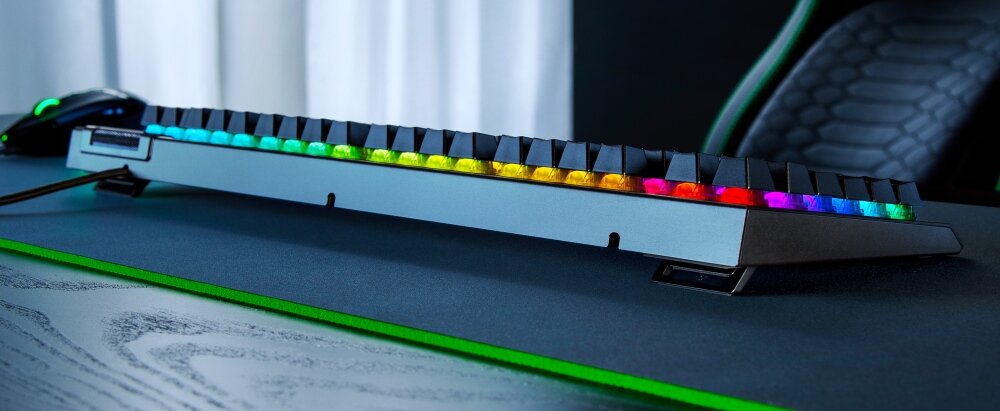 Klawiatura z podświetleniem RGB, ustawiona na biurku z podkładką. W tle widoczny jest fragment krzesła gamingowego.