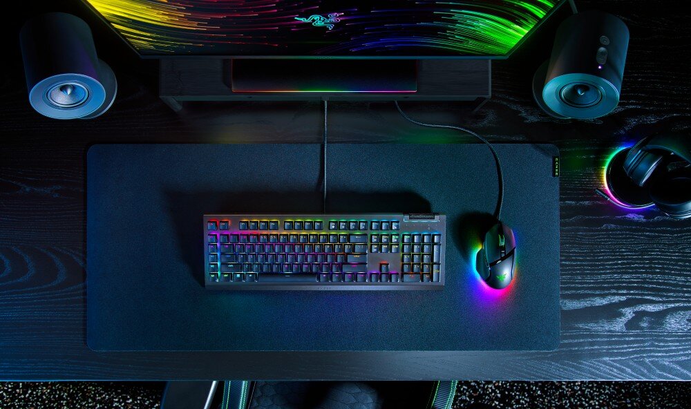 Na biurku znajduje się klawiatura z podświetleniem RGB oraz myszka, również z podświetleniem. W tle widoczny jest monitor z kolorowym wzorem oraz dwa głośniki po bokach.