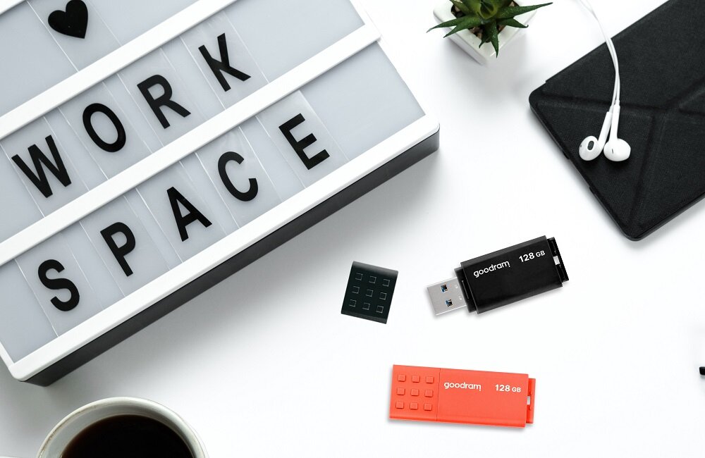 Na białym tle znajduje się czarny pendrive z napisem \'goodram 128 GB\' oraz pomarańczowy pendrive z tym samym napisem. Obok widoczna jest tablica z napisem \'WORK SPACE\', słuchawki, filiżanka kawy i roślina w doniczce.