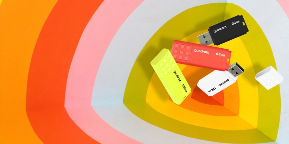 Na obrazie widoczne są cztery pendrive\'y w różnych kolorach: czarny, czerwony, żółty i biały, z logo \'goodram\' oraz oznaczeniami pojemności. Tło składa się z koncentrycznych, kolorowych pierścieni.