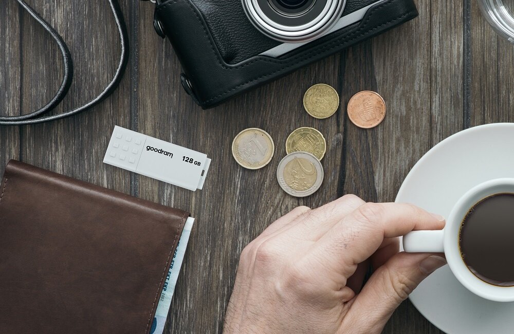Na drewnianym stole leży biały pendrive z napisem \'goodram 128 GB\', obok znajduje się aparat fotograficzny, kilka monet, brązowy portfel oraz filiżanka z kawą trzymana przez rękę.