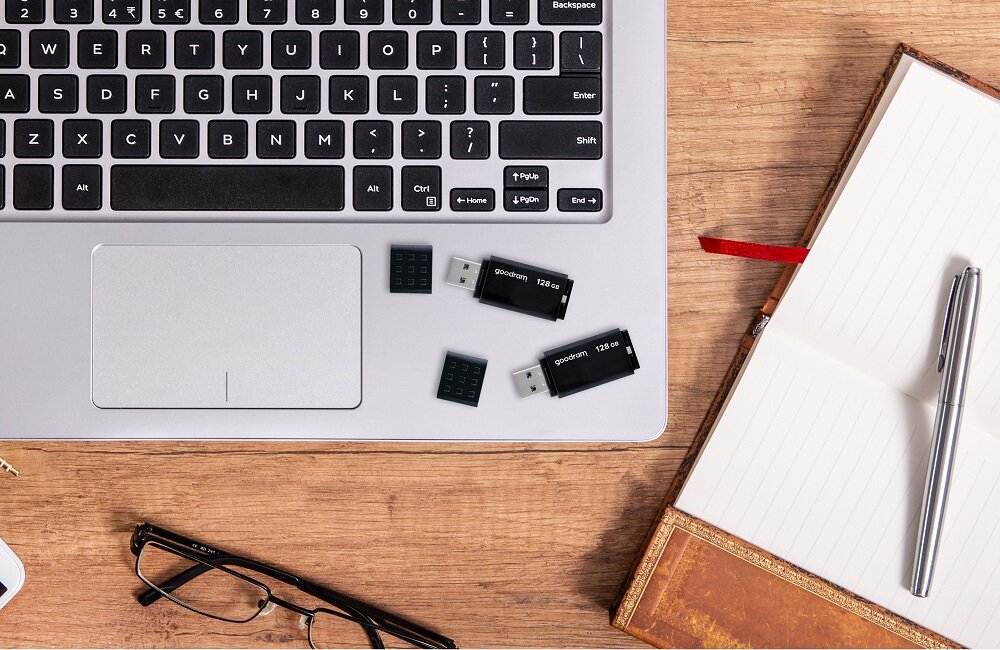 Na drewnianym blacie znajduje się otwarty laptop z widoczną klawiaturą, obok którego leżą dwa czarne pendrive\'y z napisem \'goodram 128 GB\'. Obok laptopa leży otwarty notes z długopisem oraz okulary.