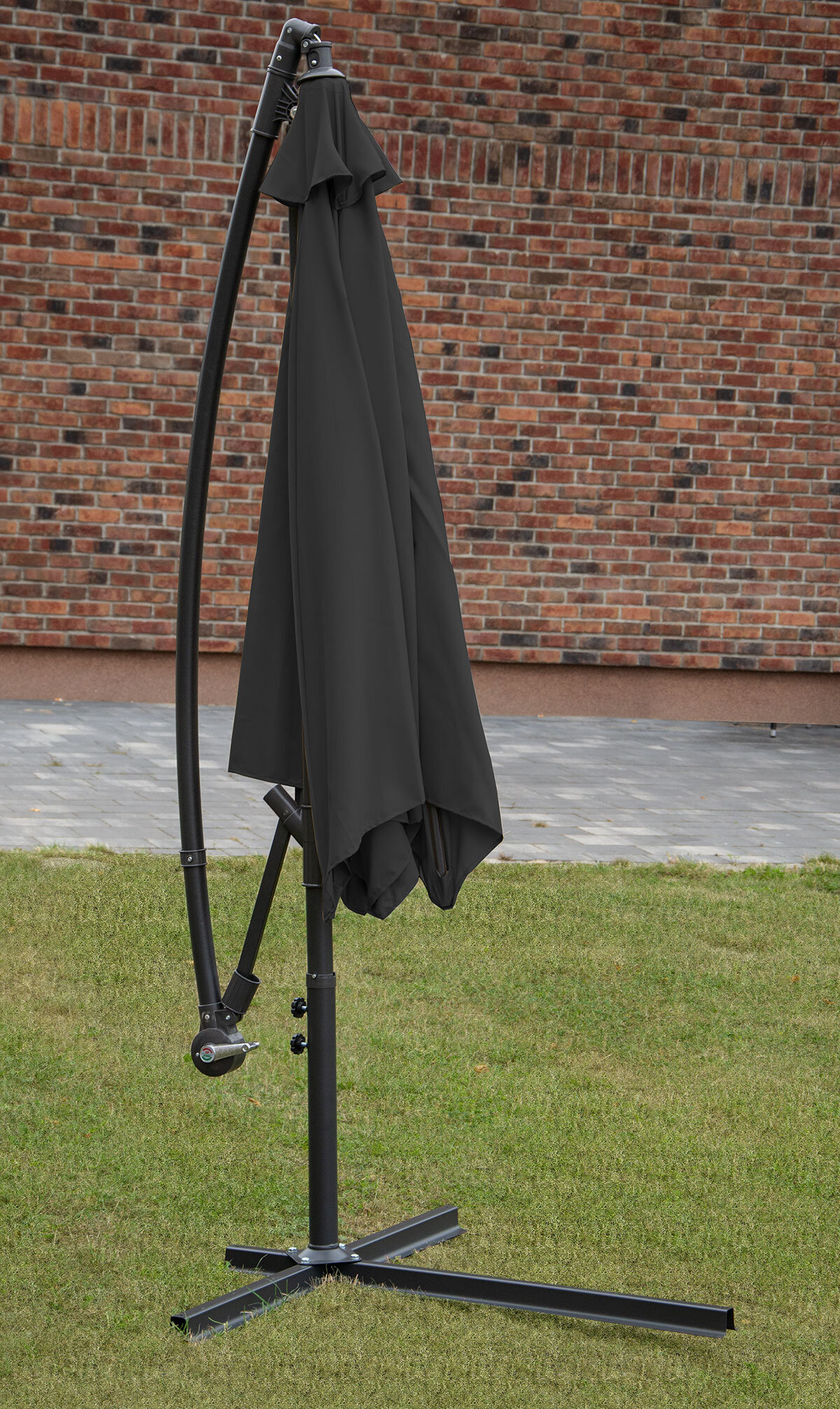 Na trawniku stoi złożony parasol ogrodowy w kolorze szarym, zamontowany na metalowym stelażu z podstawą w kształcie krzyża. W tle widoczna jest ceglana ściana budynku.