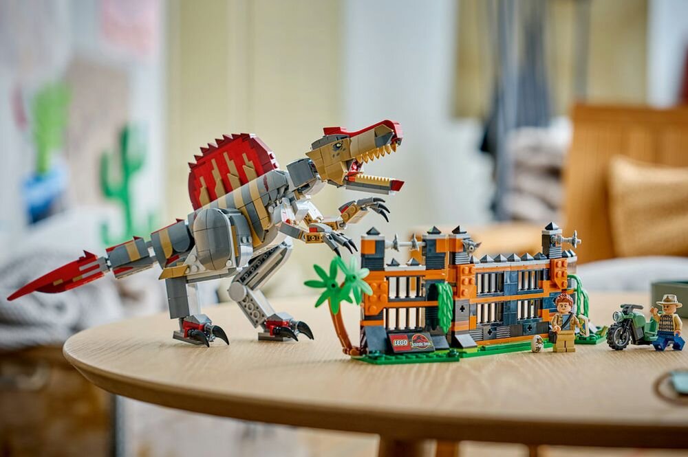 Na okrągłym stole znajduje się zestaw LEGO przedstawiający dużego dinozaura oraz fragment ogrodzenia z palmą i figurkami. Widoczne jest logo LEGO Jurassic World na ogrodzeniu.