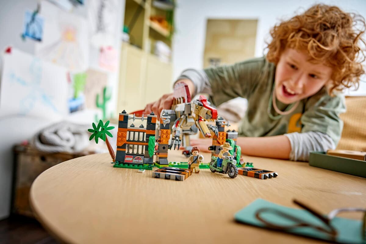 Na stole znajduje się zestaw klocków LEGO przedstawiający scenę z dinozaurem i budynkiem. W tle widać dziecko bawiące się zestawem.