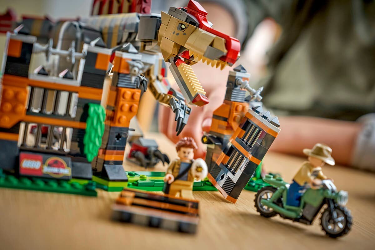 Zestaw klocków przedstawia scenę z dinozaurem i dwoma figurkami ludzików, z których jeden jest na motocyklu. W tle widoczne są elementy konstrukcji z logo LEGO i Jurassic World.