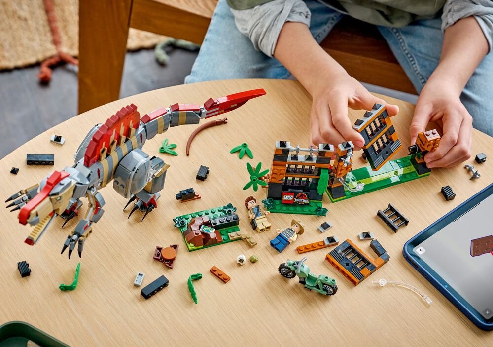 Na okrągłym stole znajdują się klocki LEGO przedstawiające dinozaura, budynki i postacie. Dziecko bawi się zestawem, a obok leży tablet.
