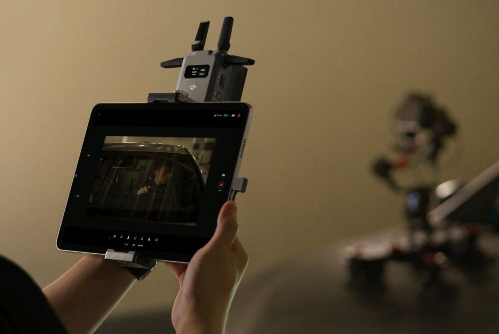 Osoba trzyma tablet z zamontowanym urządzeniem nad ekranem, na którym wyświetlany jest obraz z filmu. Na urządzeniu widoczne jest logo DJI.