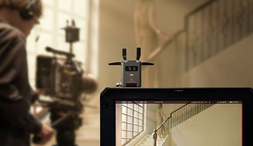 Na obrazie widać urządzenie z logo DJI umieszczone na monitorze, na którym wyświetlany jest obraz wnętrza z klatką schodową. W tle znajduje się osoba obsługująca kamerę filmową.