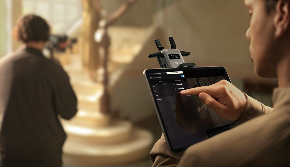 Osoba obsługuje tablet z zamontowanym urządzeniem, na ekranie widoczny jest interfejs aplikacji. W tle znajduje się inna osoba z kamerą, a otoczenie jest rozmyte.