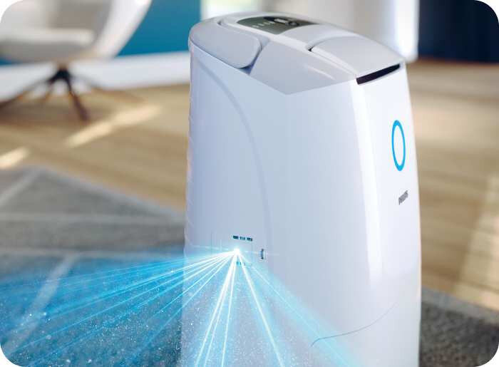 Na obrazie widoczny jest biały osuszacz powietrza z niebieskim podświetleniem i logo PHILIPS. Urządzenie stoi na dywanie, a z jego boku emitowane są niebieskie promienie świetlne.