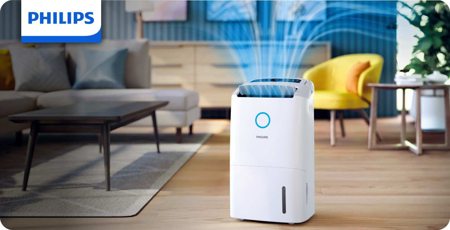 Na obrazie znajduje się biały osuszacz powietrza z logo PHILIPS, ustawiony w nowoczesnym salonie. Z urządzenia wydobywają się niebieskie linie symbolizujące przepływ powietrza.