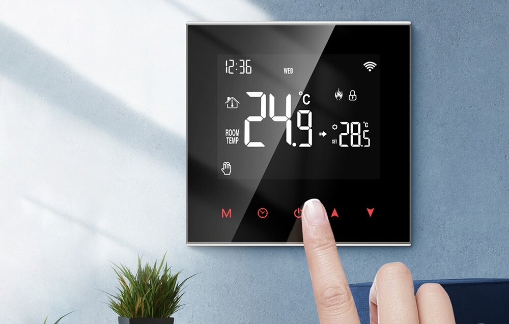Na ścianie zamontowany jest nowoczesny termostat z wyświetlaczem pokazującym temperaturę 24,9°C oraz inne ikony i informacje, takie jak godzina 12:36 i dzień tygodnia \'WED\'. Obok termostatu widoczna jest ręka wskazująca na przycisk dotykowy.